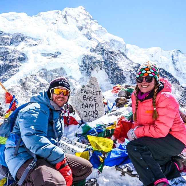 A couple at Everest Base Camp 2019 stone surrounded by prayer flags and a snow capped mountain in the background