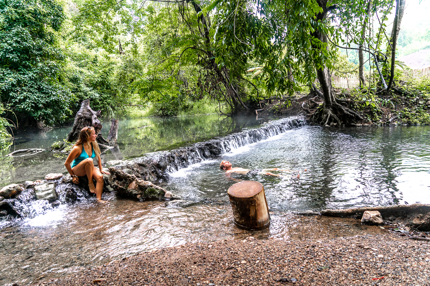 A couple relaxing at hot springs in the forest - girl on the edge, boy floating in the water