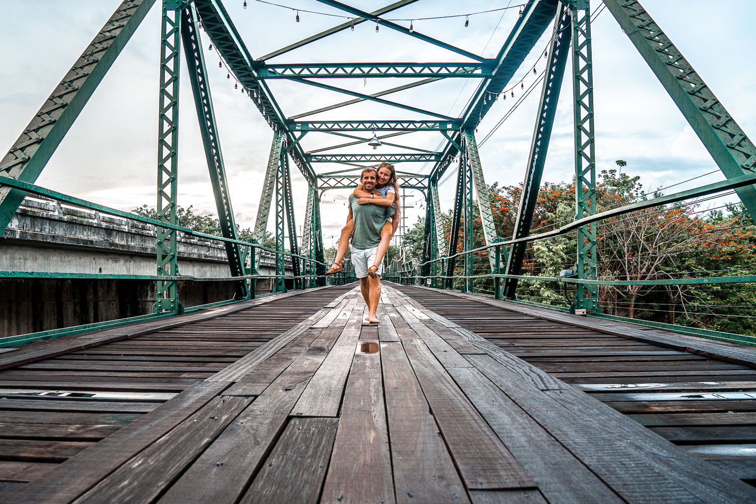 A man carrying a woman piggy back over a metal bridge with dark wooden plank floor