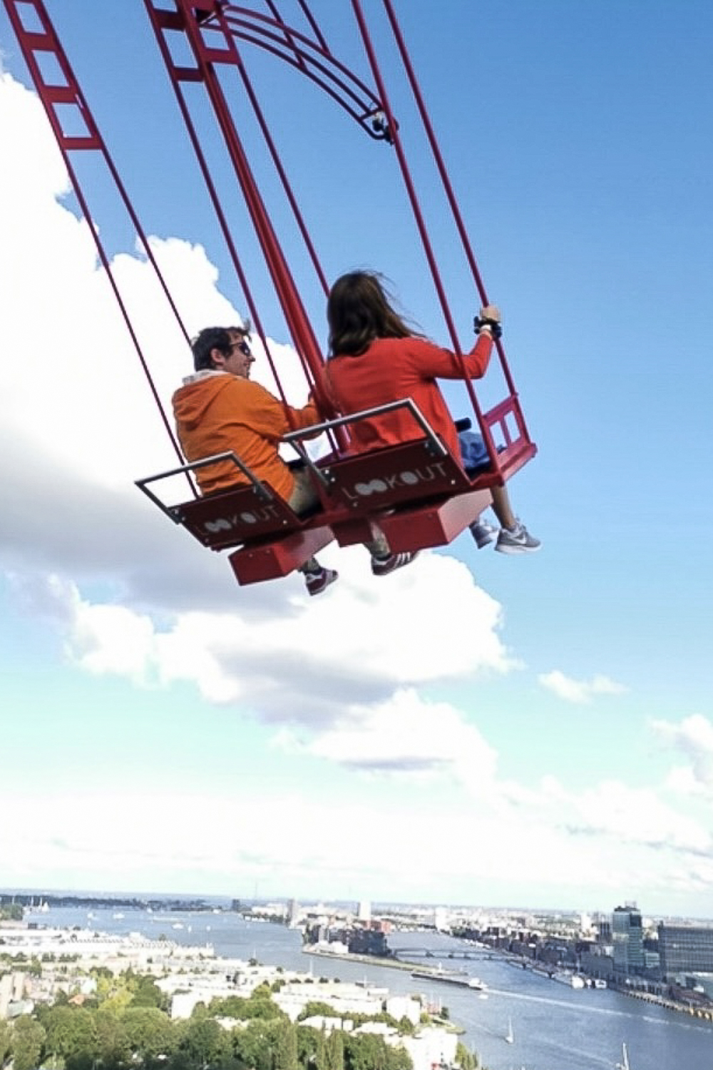 Two people on a large seat swing, swinging very high above a wharf in Amsterdam