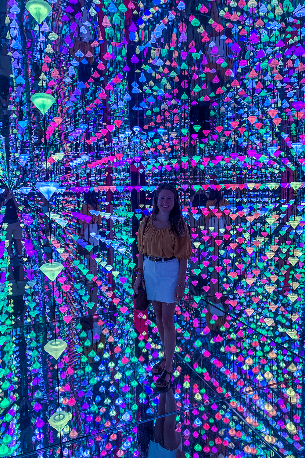 Girl stood in an art exhibit of countless blue, green, and pink lights suspended on strings from the ceiling