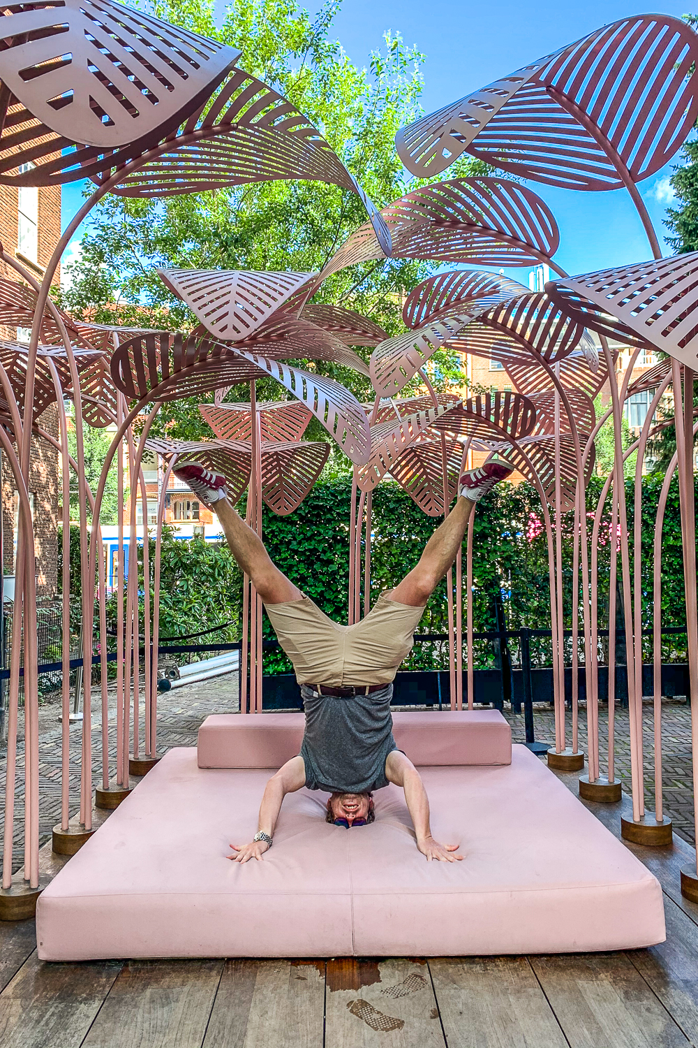 Man doing a headstand on a rose pink bed with large rose pink metal leaf structures above