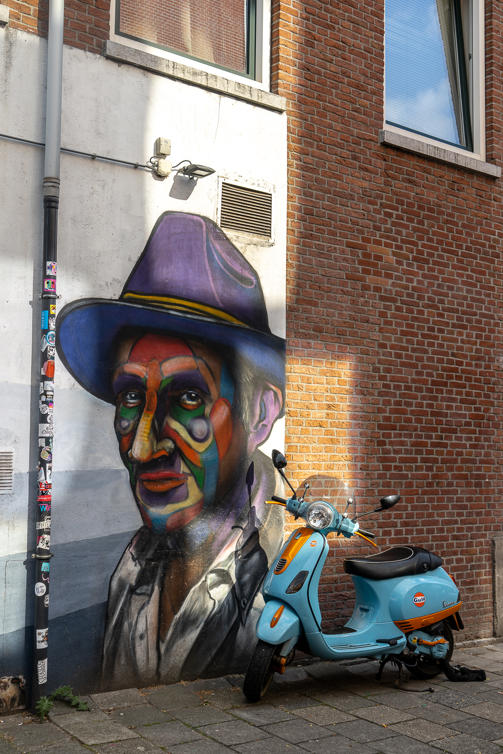 Street art of a man wearing a fedora - Cool blue and orange Vespa chained up nearby