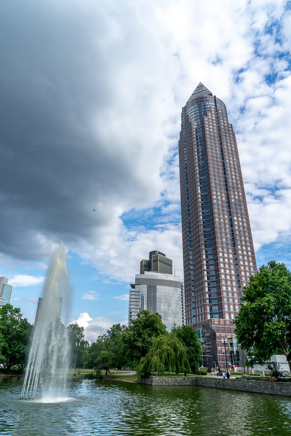 A tall skyscraper with a pointed roof and a large lake and fountain in front