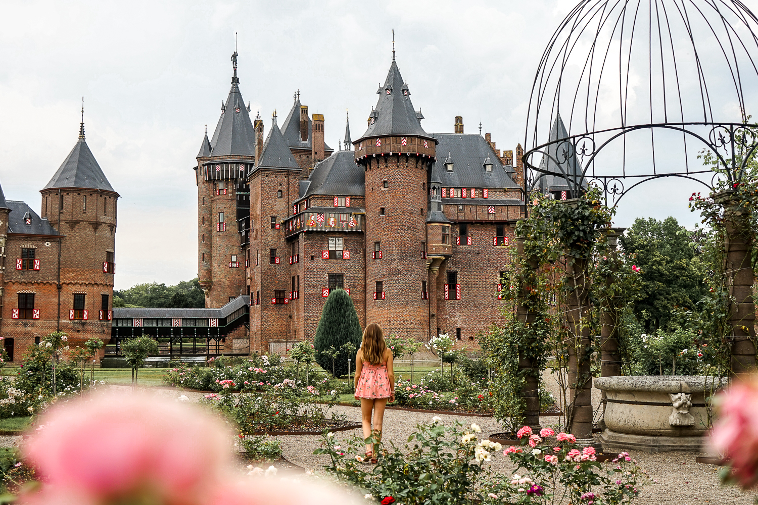 Girl in a pink dress standing in the rose garden of a huge red castle. De Haar Castle in Netherlands). Framed by pink roses.
