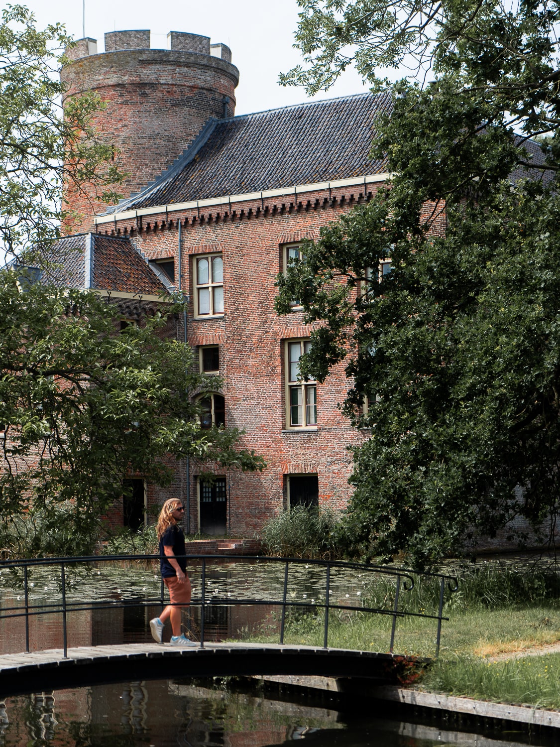 A red brick castle with a small bridge with a man standing on it set amongst trees