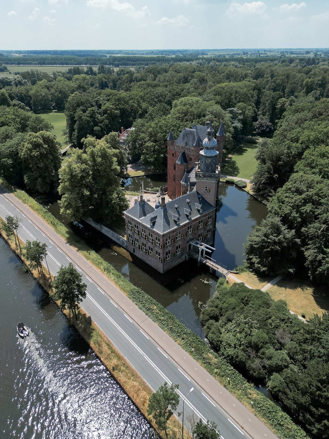 A high aerial shot of a red brick Dutch castle surrounded by a moat and trees next to a lake with a boat