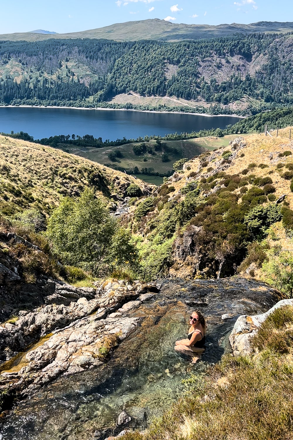 A woman sat in a small pool at the top of a waterfall overlooking a lake