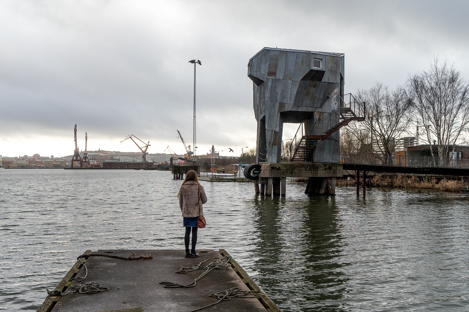 A girl stood on the edge of a man-made lake, part of the dock area of Gothenburg. With an industrial looking building on thick stilt like supports
