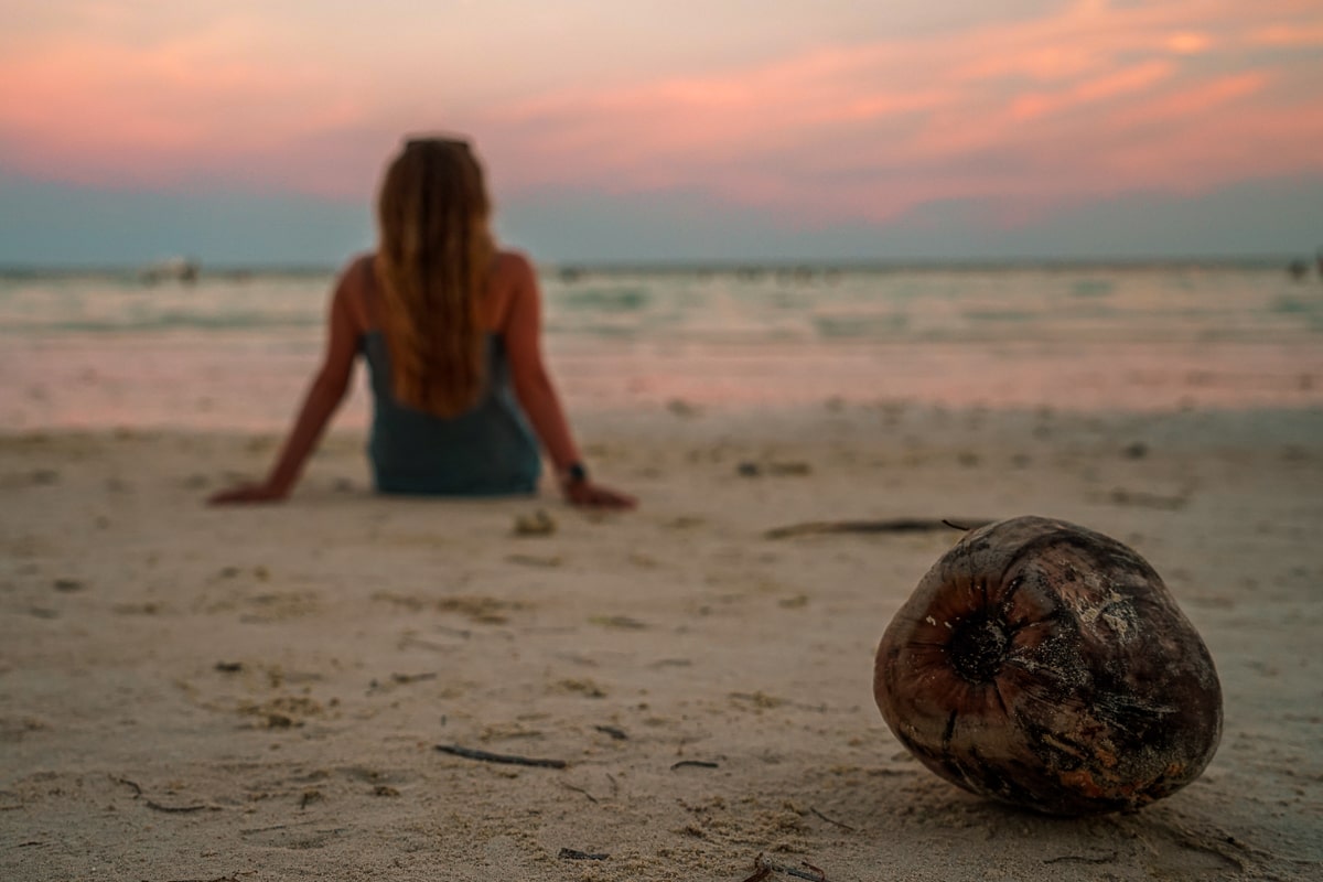 A close up of a coconut on a beach with a girl sat behind
