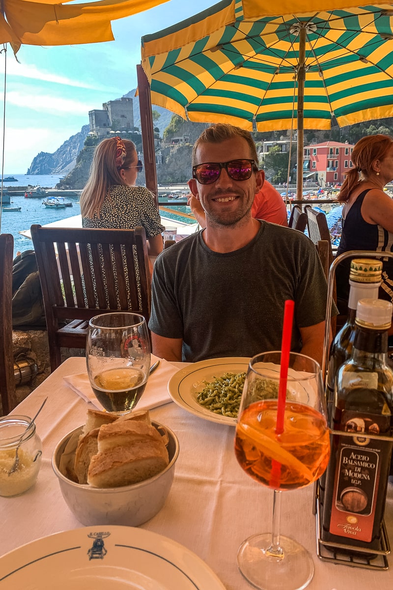 A man at an al fresco restaurant with yellow and green parasols. A plate of pesto pasta, beer, and aperol spritz on the table