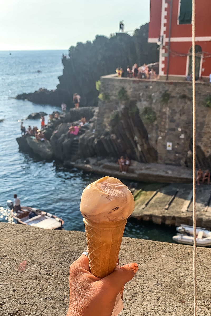 A woman holding an ice cream cone with a red building and the sea behind