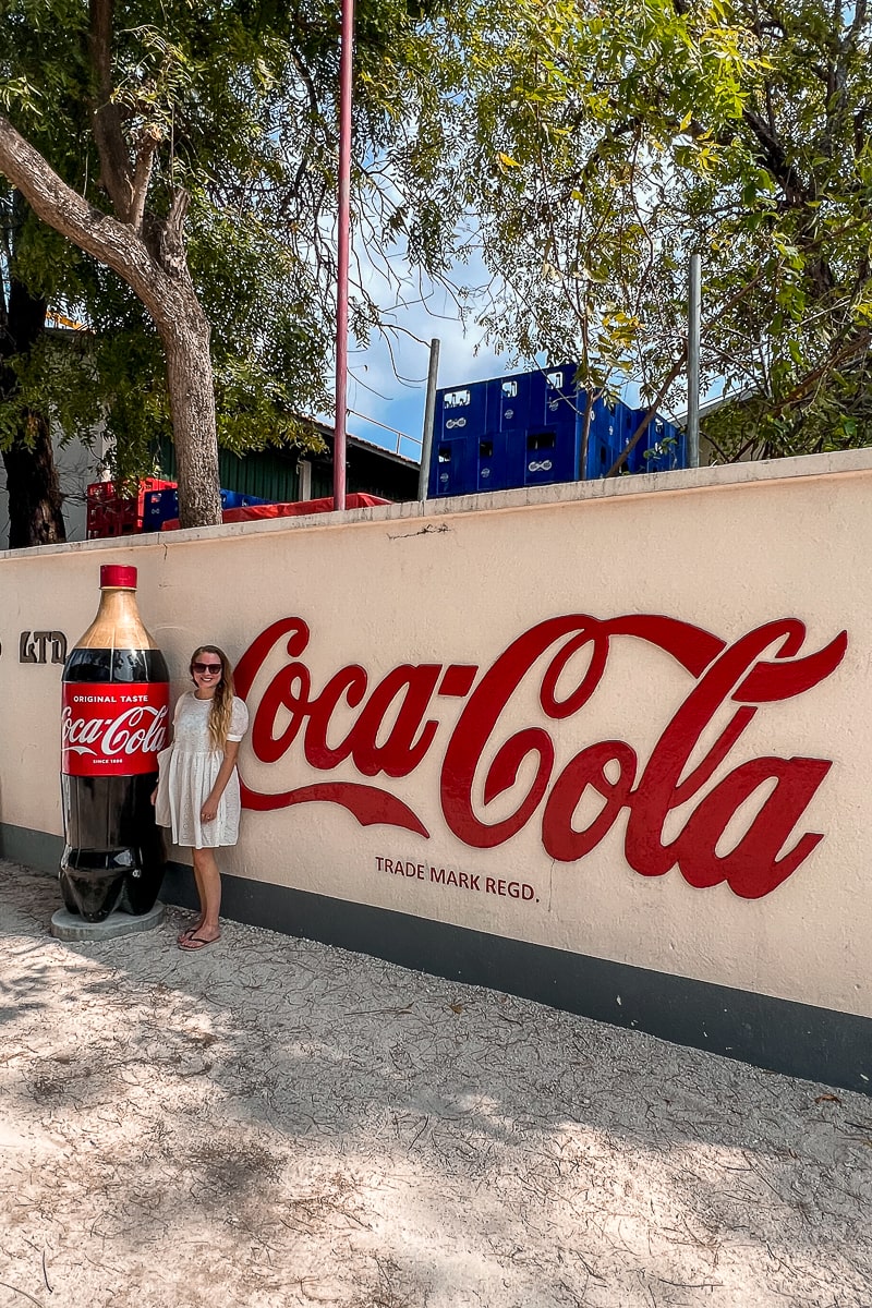 A person stood next to a human-sized Coca-cola bottle and Coca-cola sign of the Thulusdhoo factory