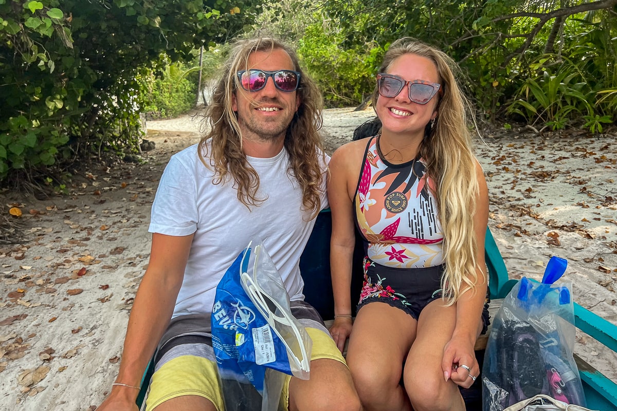 A tanned couple in beachwear smile as they're transported on the back of a small minivan along a sandy trail