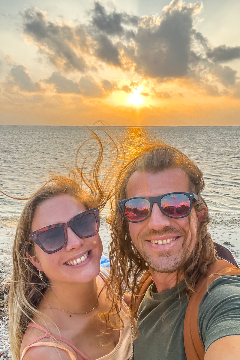 A couple smiling with shades on as the sun sets over the ocean behind them