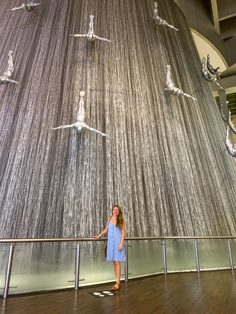 A girl standing in front of a fountain with statues of men diving down it