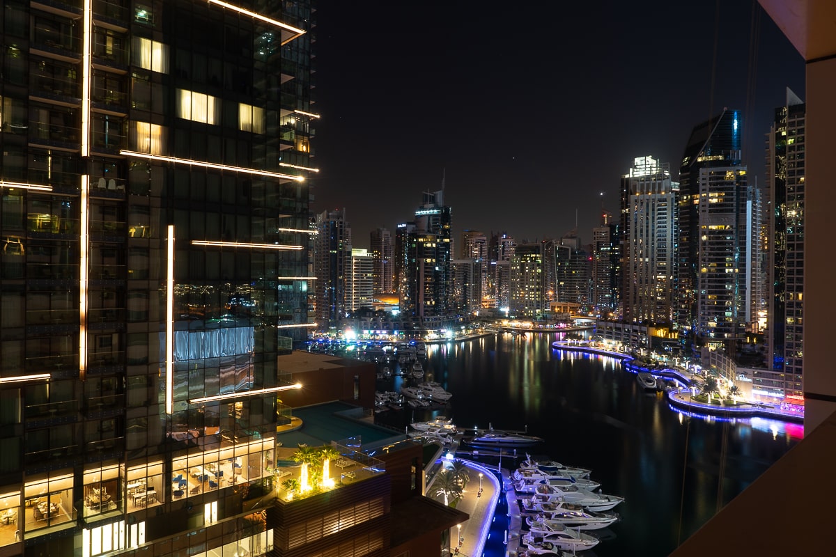 A night photo of Dubai Marina. Many skyscrapers surround the water which is filled with docked boats and yachts