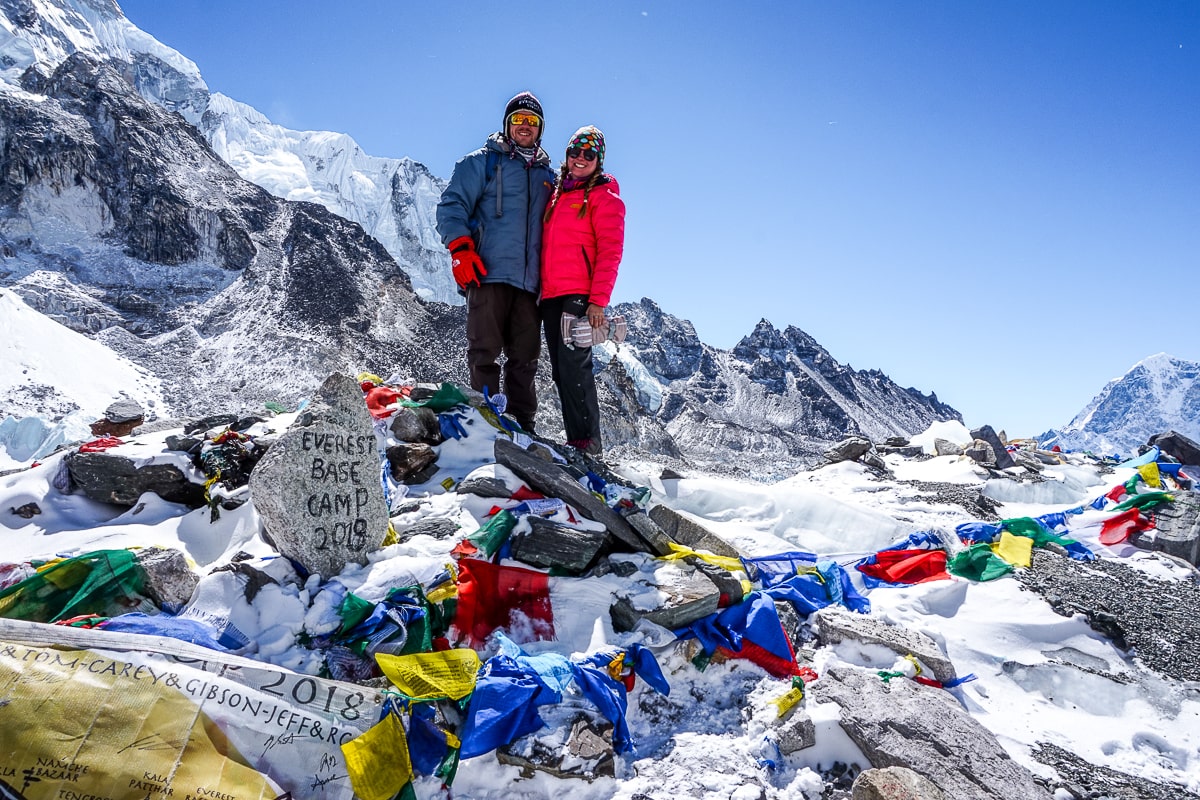 A couple stood next to the "Everest Base Camp 2019" stone surrounded by prayer flags and snow covered mountains