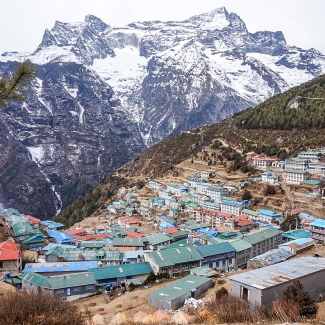 A wide view of Namche Bazaar from above. Metal coloured roofs, evergreen trees, and huge snowy mountains