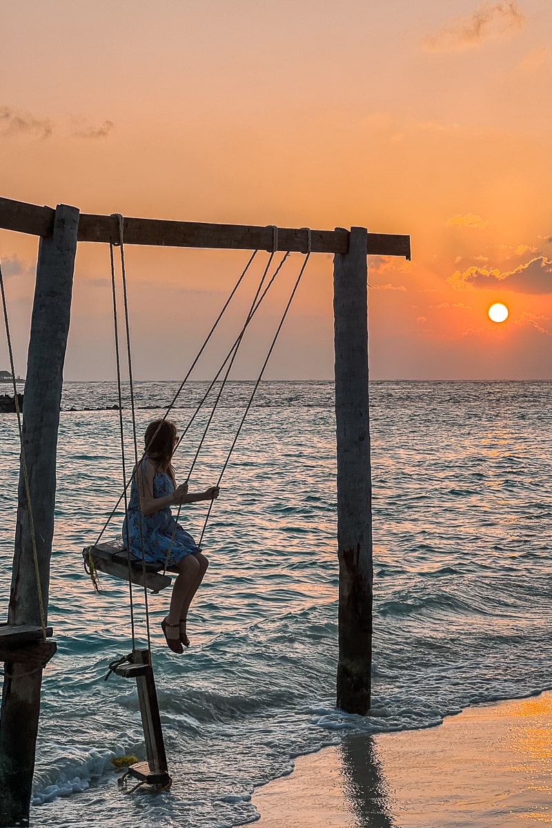 A person swinging on a wooden swing on the edge of the ocean watching the sun going down