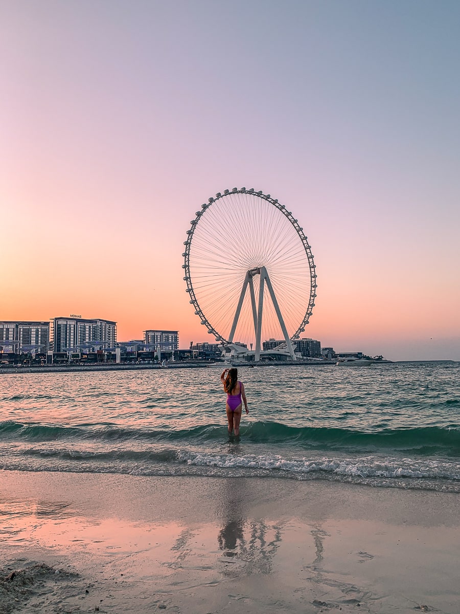 A girl wading in the sea at sunset with a big ferris wheel behind. The sky burns orange and purple