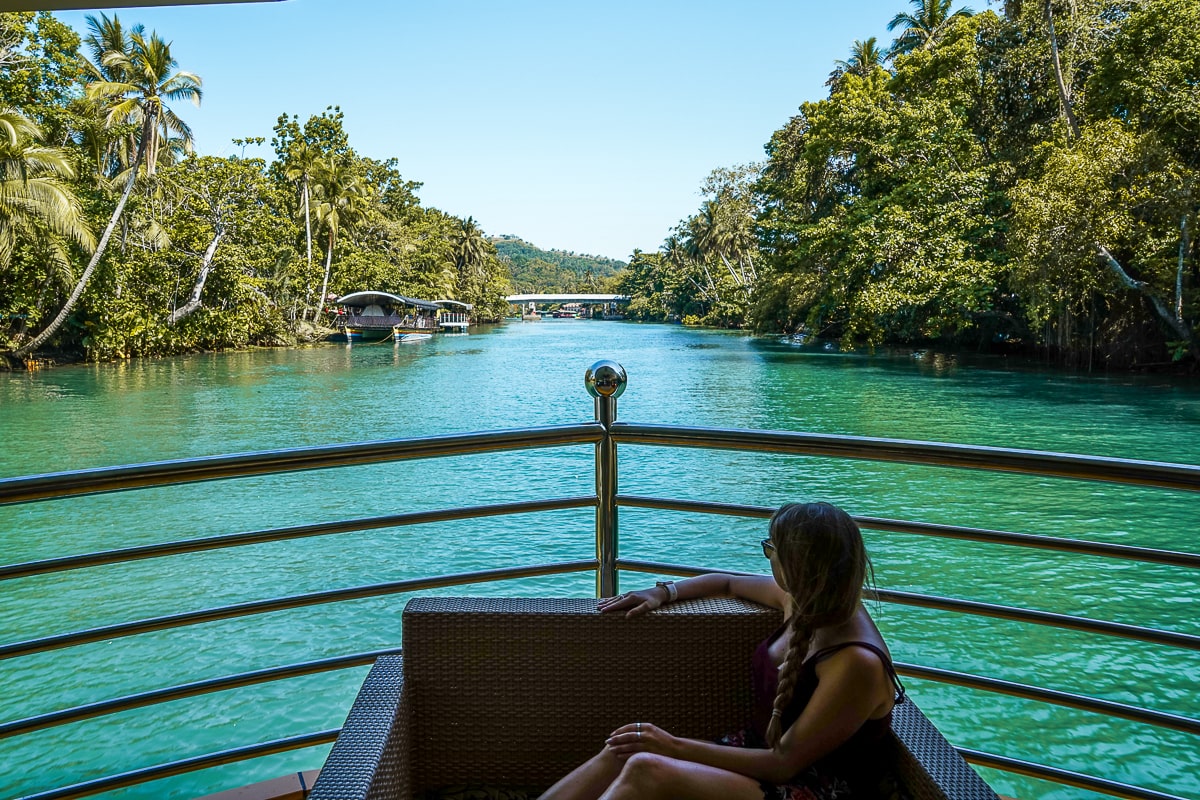 A woman sat on an armchair at the bow of a large boat sailing down a jungle river