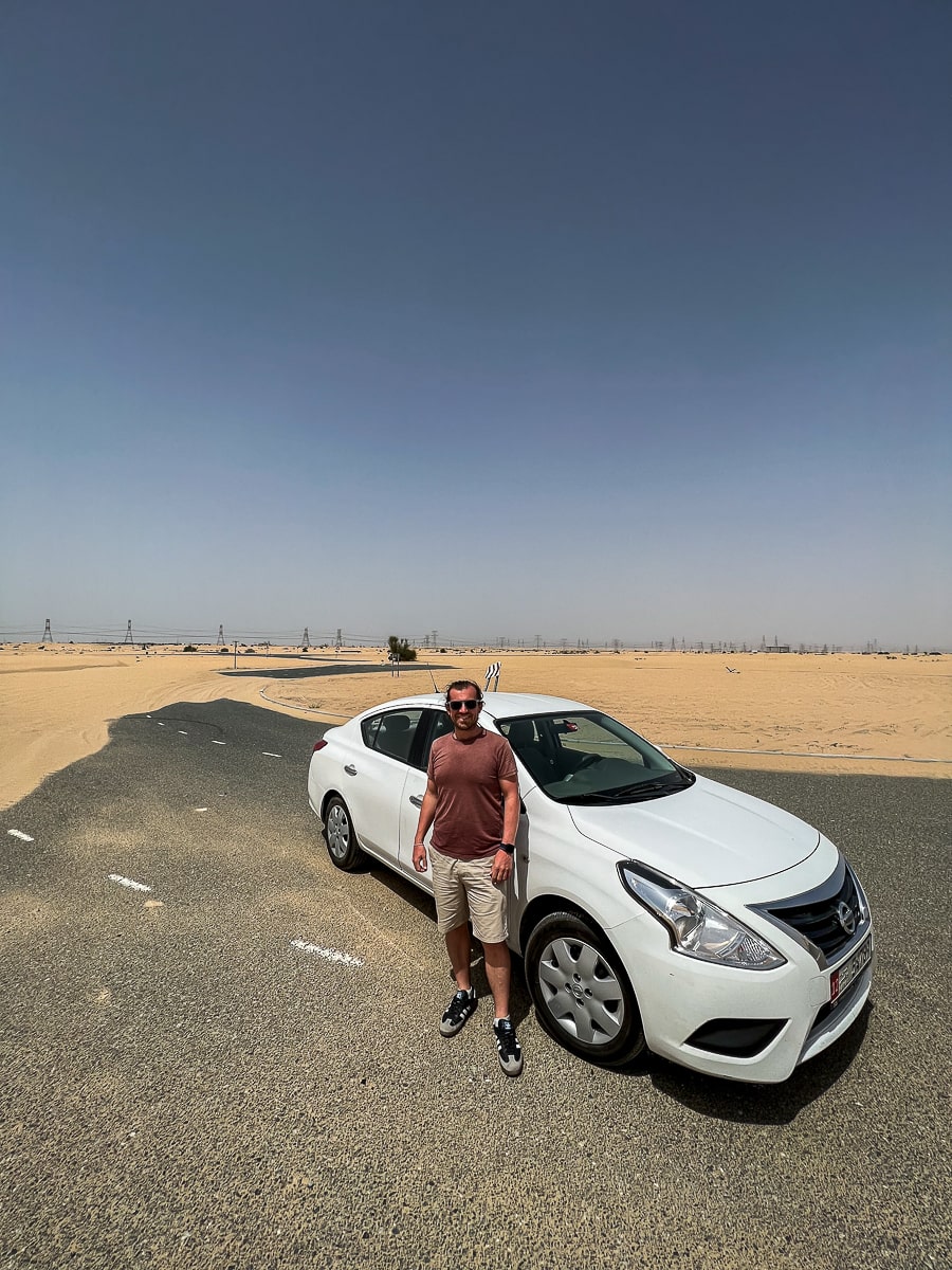 A man standing in front of a white car in the desert. The sand is covering the road behind
