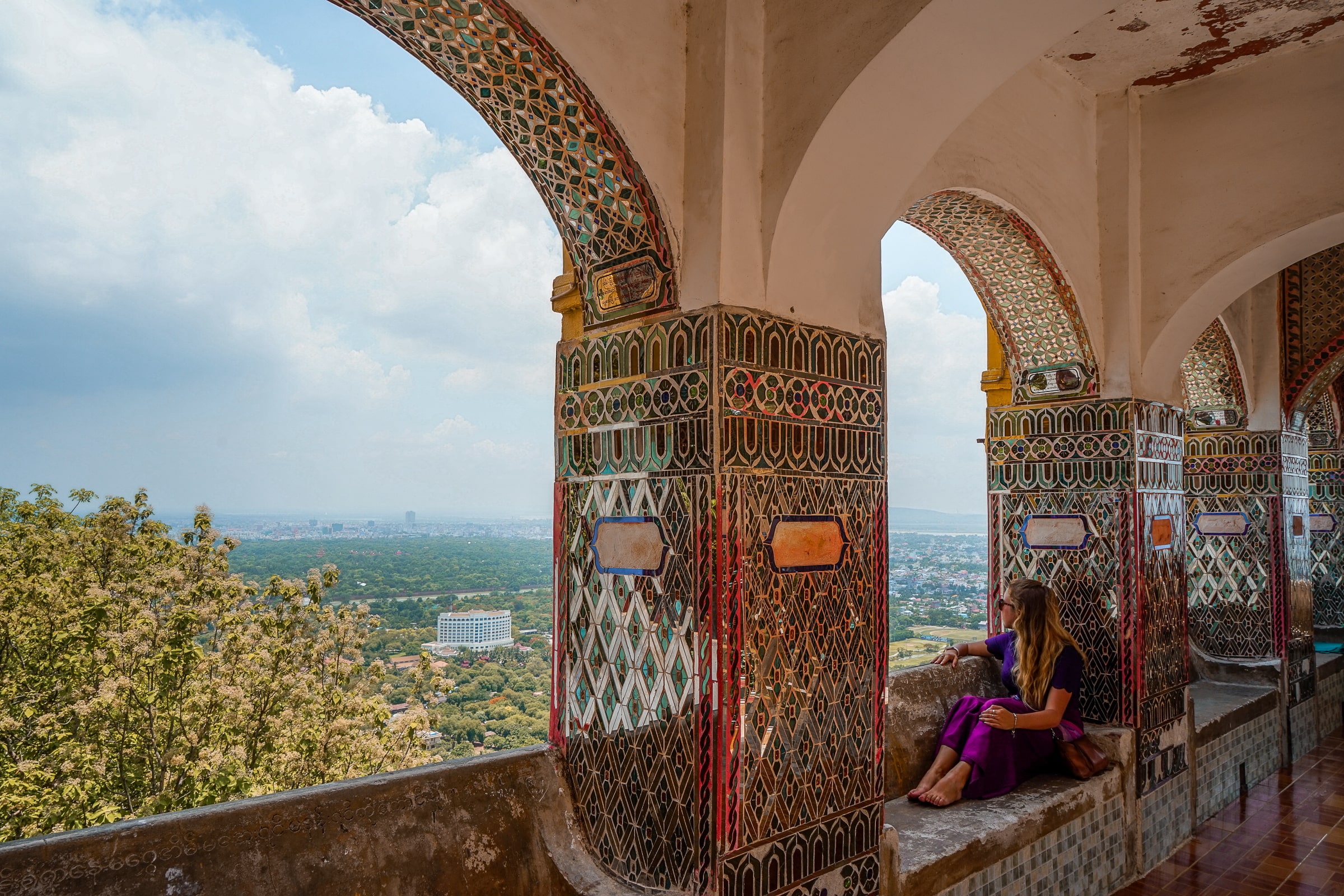 A woman sitting on a balcony decorated with gemstones and tiles looking out over a high viewpoint