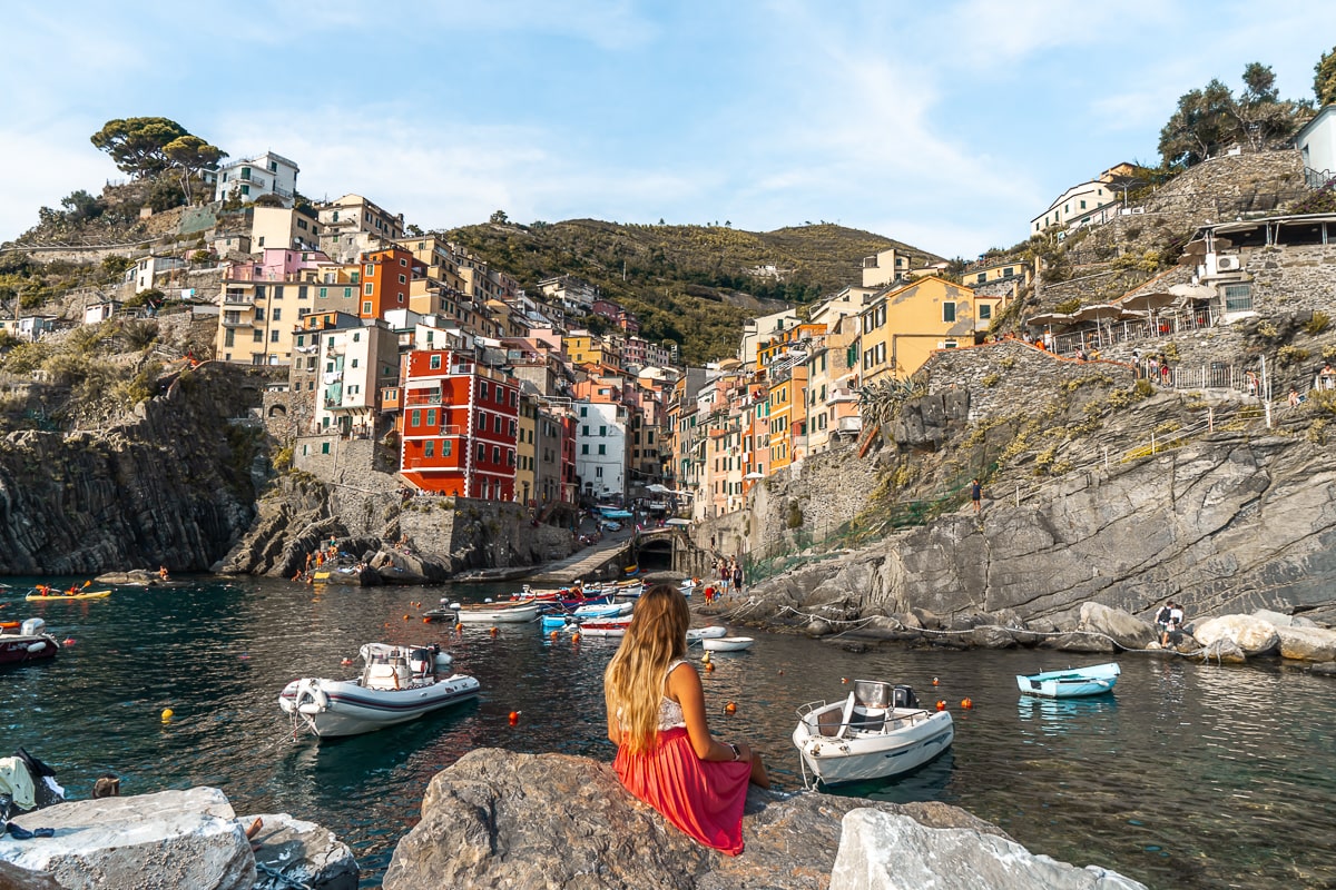 A woman sat on some rocks in the sea with the colorful town of Riomaggiore behind and many small boats anchored in the water