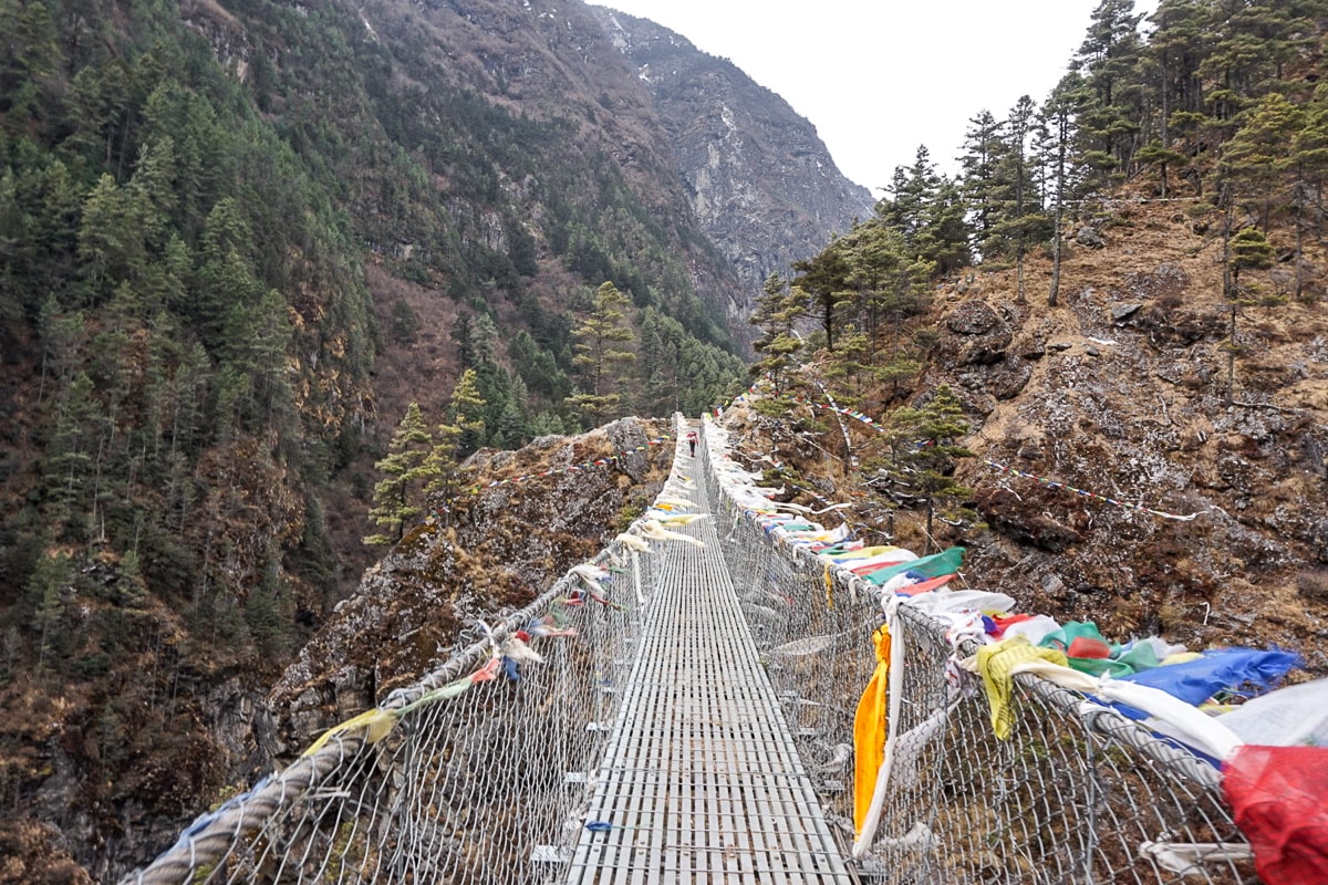 A metal suspension bridge draped in prayer flags, spanning a deep valley