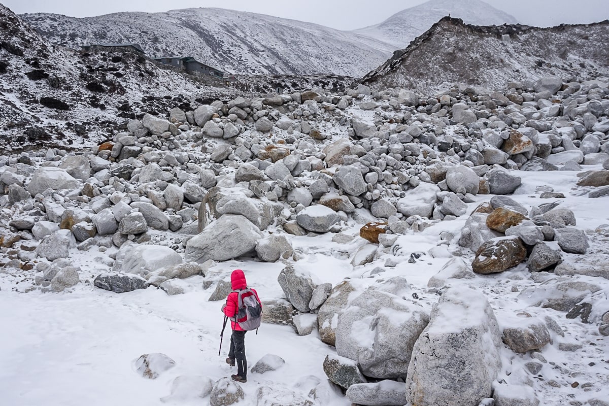 A woman in a pink winter jacket with trekking poles traversing a snowy valley covered in boulders