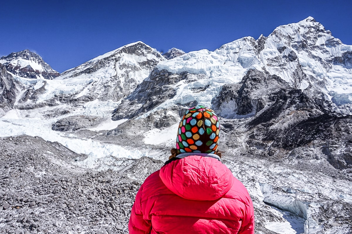A woman in a spotty, colorful beanie hat and pink winter jacket overlooking a glacier and Everest