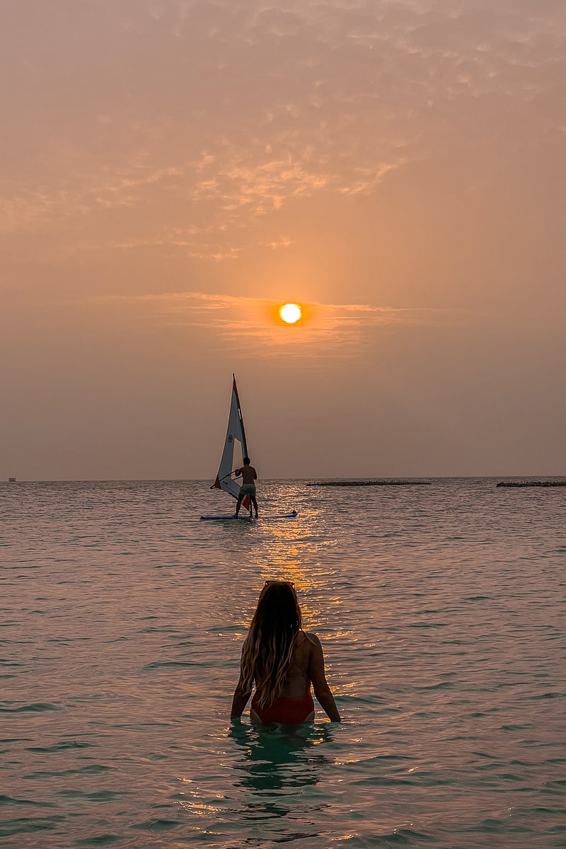 A person wading in the ocean watching a windsurfer in the distance