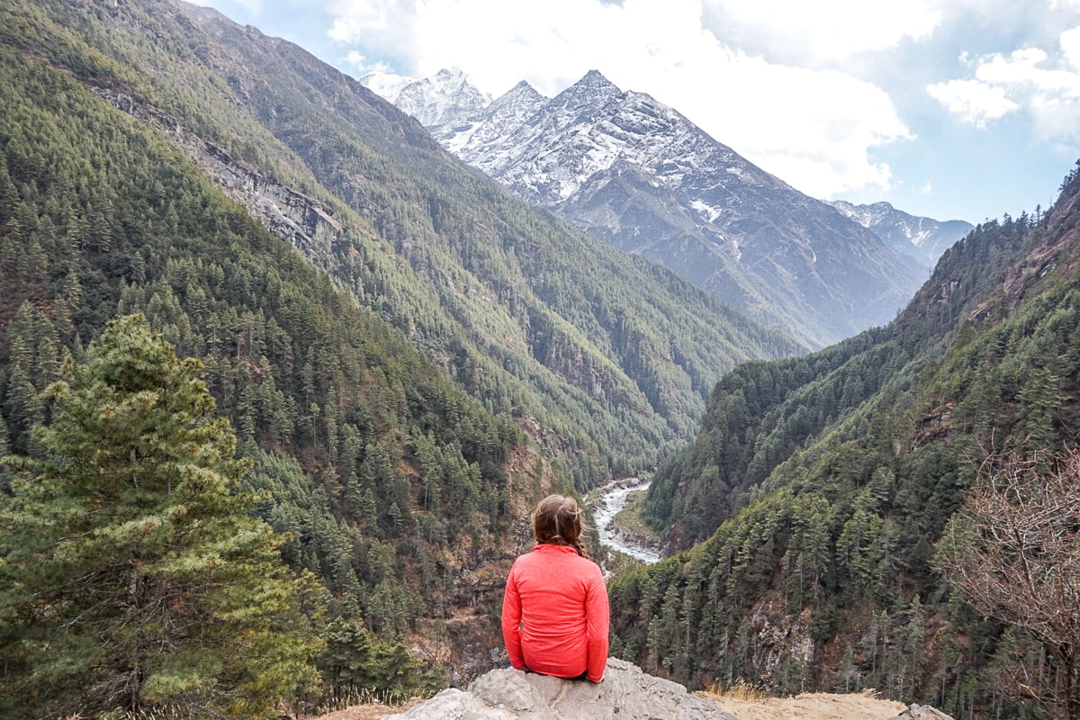 A woman sat on a rock with a view over a deep ravine with a river and evergreen trees