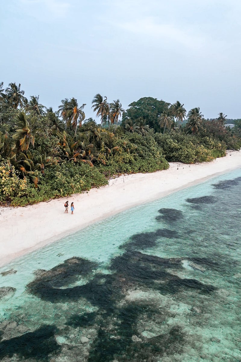 A drone shot of a couple walking along a deserted, tropical beach with palm trees and a shallow clear sea with seagrass and rocks