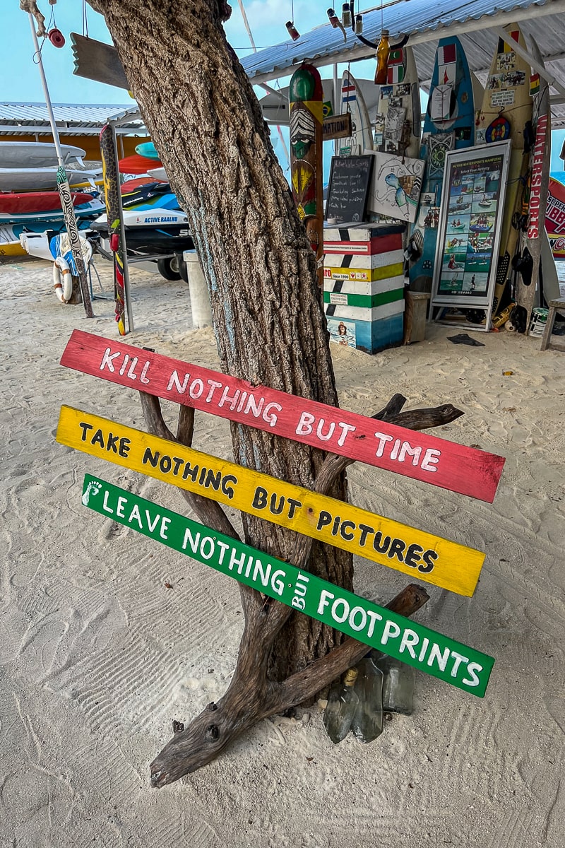A set of wooden signs pinned to a tree at a beach. From top to bottom - a red sign reading "kill nothing but time", a yellow sign reading "take nothing but pictures", and a green sign reading "leave nothing but footprints"