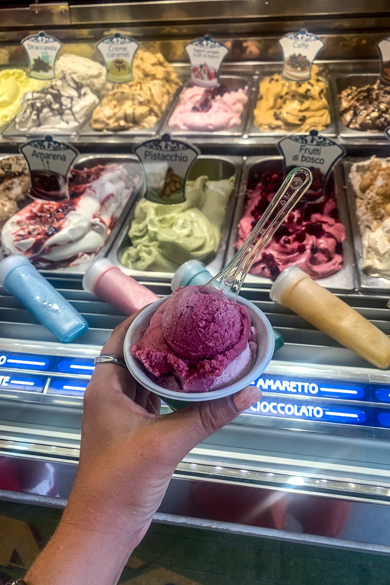 A woman holding a one scoop tub of berry gelato in front of a selection of ice creams