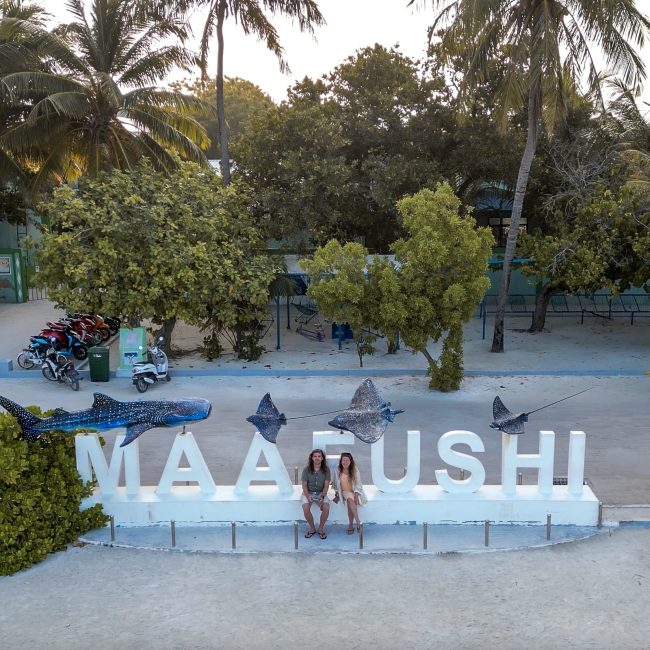A drone shot of a couple sat on a large "Maafushi" white statue sign with sculptures of marine life
