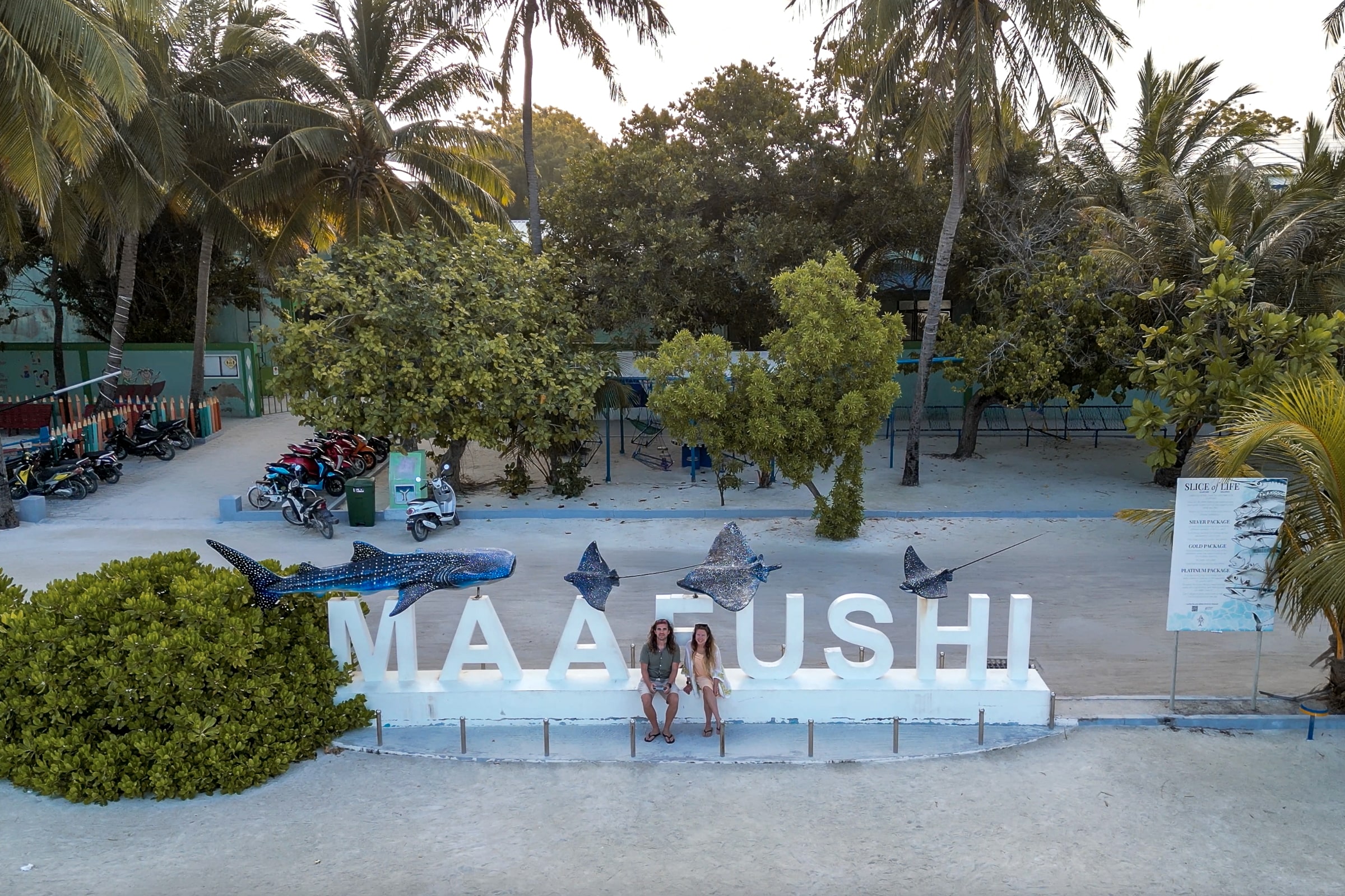 A drone shot of a couple sat on a large "Maafushi" white statue sign with sculptures of marine life
