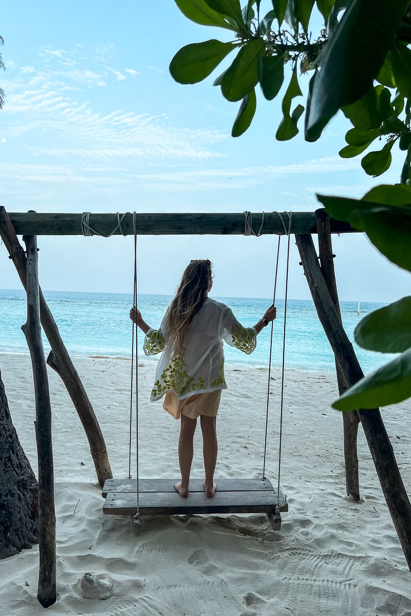 A girl stood up on a large wooden swing at a beach, looking out to the ocean