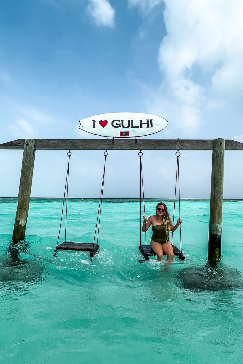 A girl sat on a swing in the sea with an "I heart Gulhi" sign on a mini surf board above