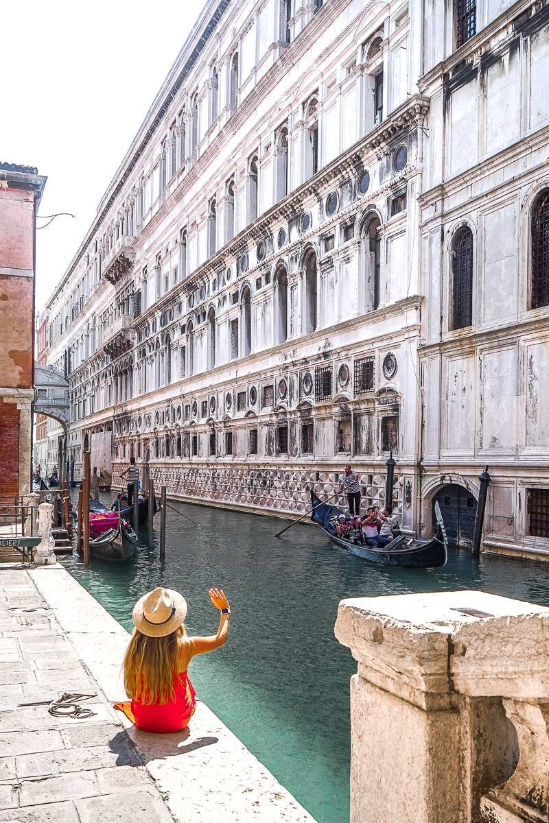 A woman in a red dress sat on the edge of the canal in Venice waving at a passing gondola