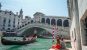 The white Rialto bridge with a gondola passing underneath and a couple sat watching