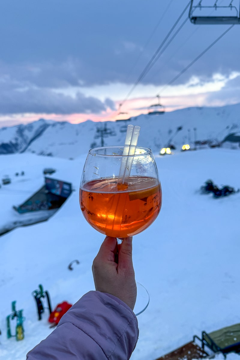 A point of view of a girl holding an aperol spritz out with snowy mountains and ski slopes in the background