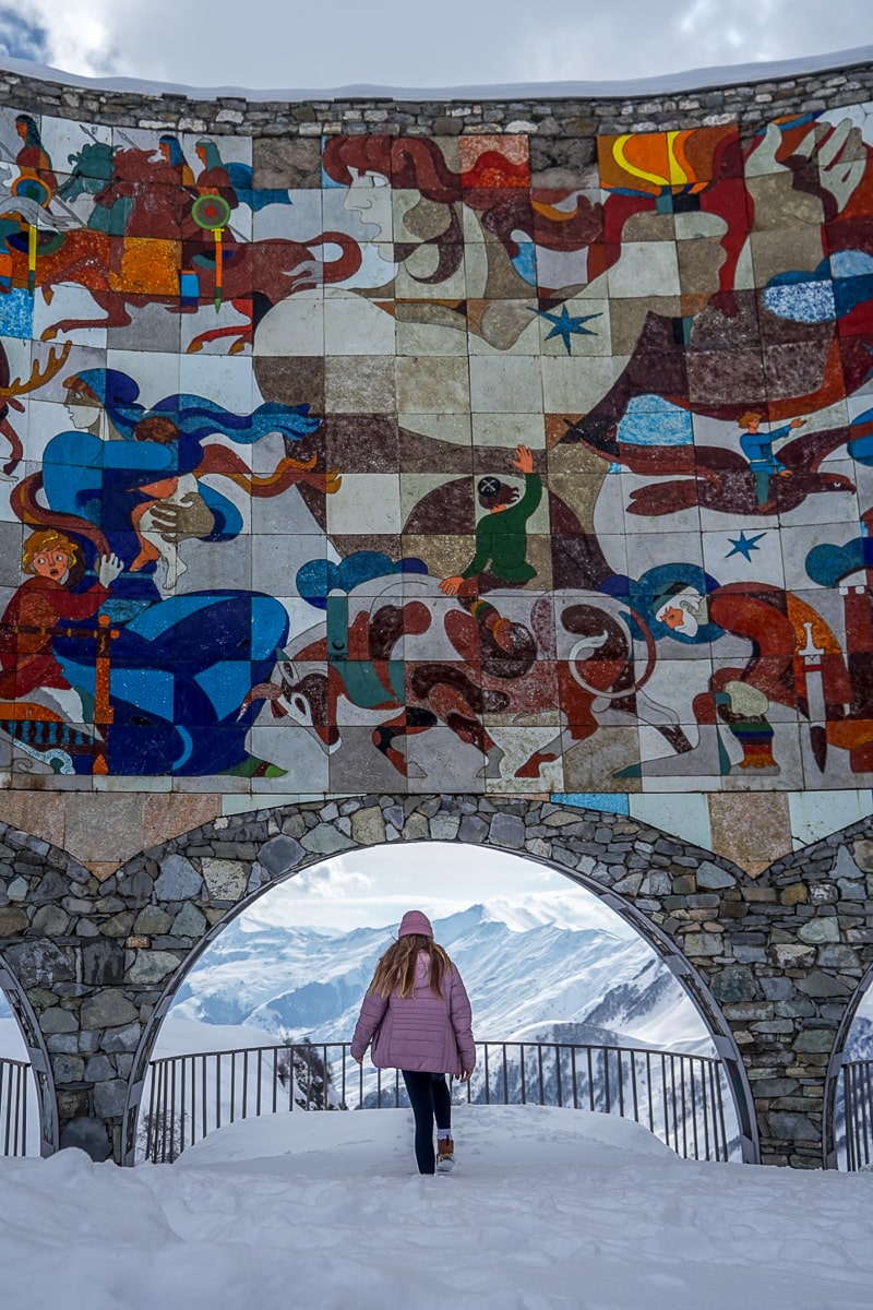 A girl stood in an archway with a view of snowy mountains in the background beneath a vibrant, abstract mosaic structure