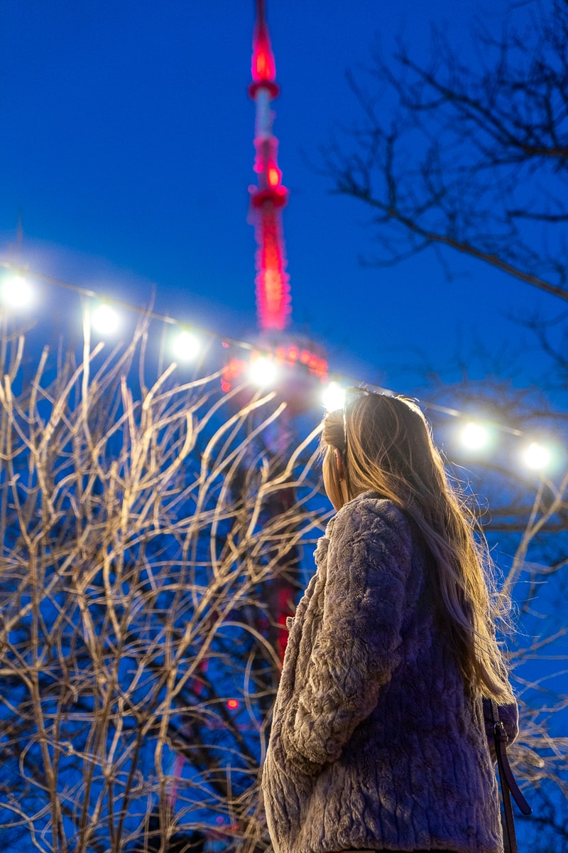 A girl looking up at the red and white TV tower in Tbilisi with fairy lights surrounding her