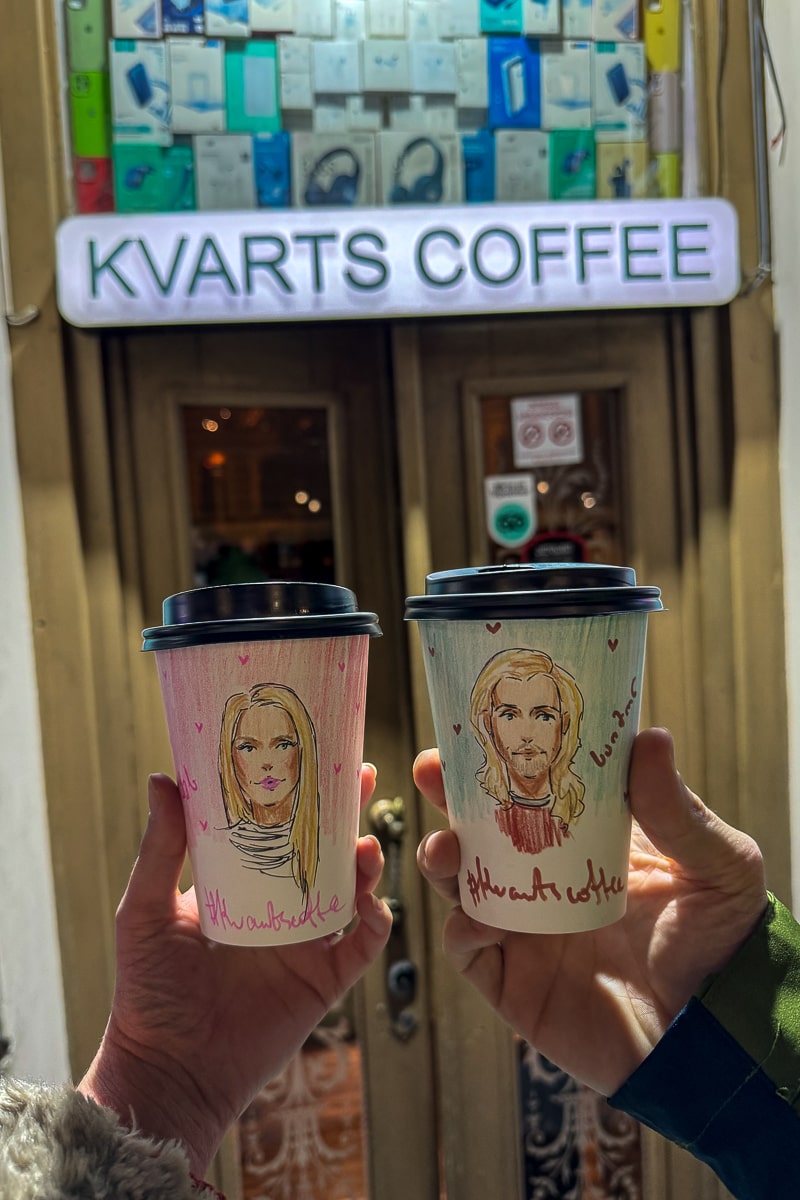People holding two paper coffee cups with portraits of a man and a woman drawn on them in front of an illuminated "Kvarts Coffee" sign