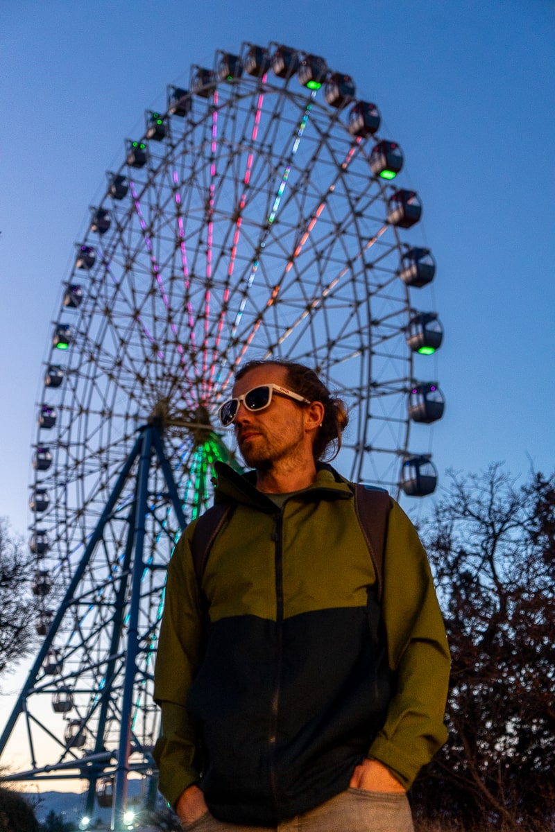 Photo of a man with sunglasses standing in front of a large ferris wheel as the sun sets
