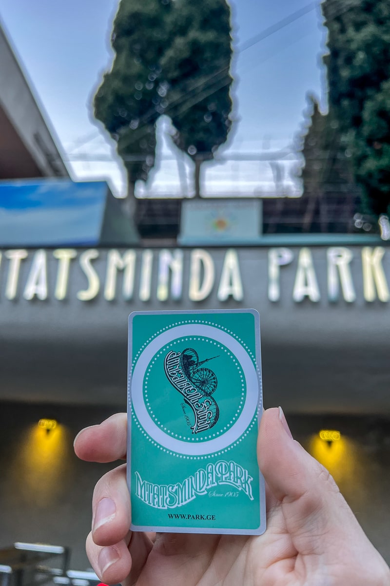 Holding a green card that reads "Mtatsminda Park" in front of an illuminated sign reading the same