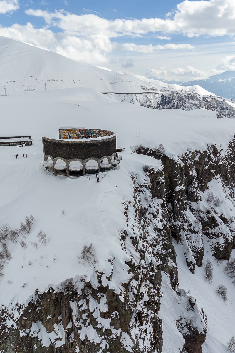 A snowy landscape of mountains and a steep cliff with a circular monument atop the hill